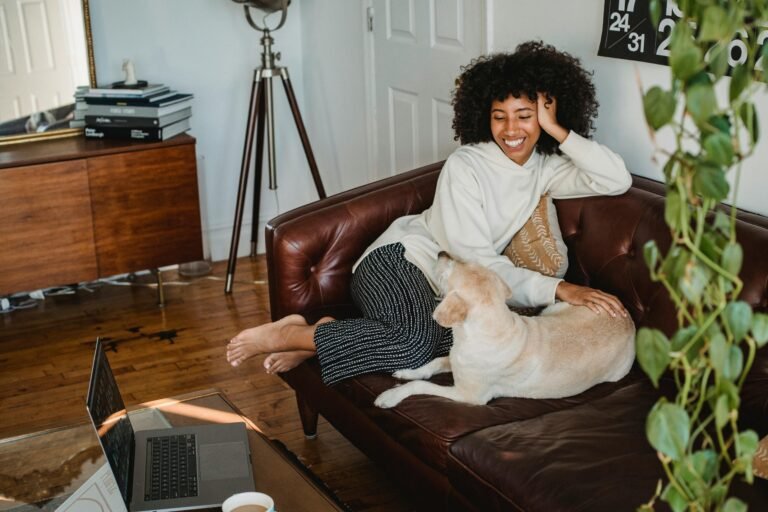 Happy young African American woman sitting on couch and resting with adorable dog with computer and cup of coffee on table in living room