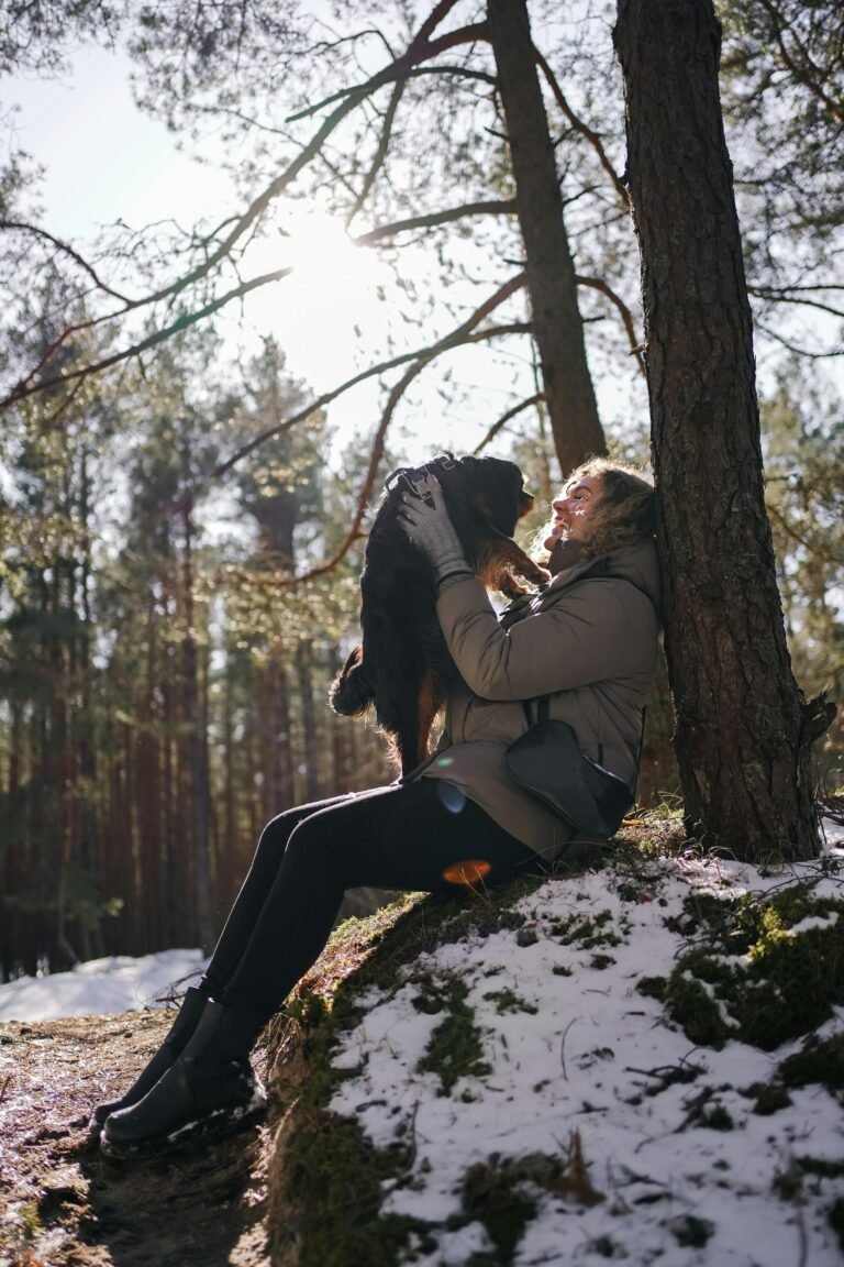 A joyful woman embraces her dog in the winter forest of Jūrmala, Latvia, basking in the warm sunlight.