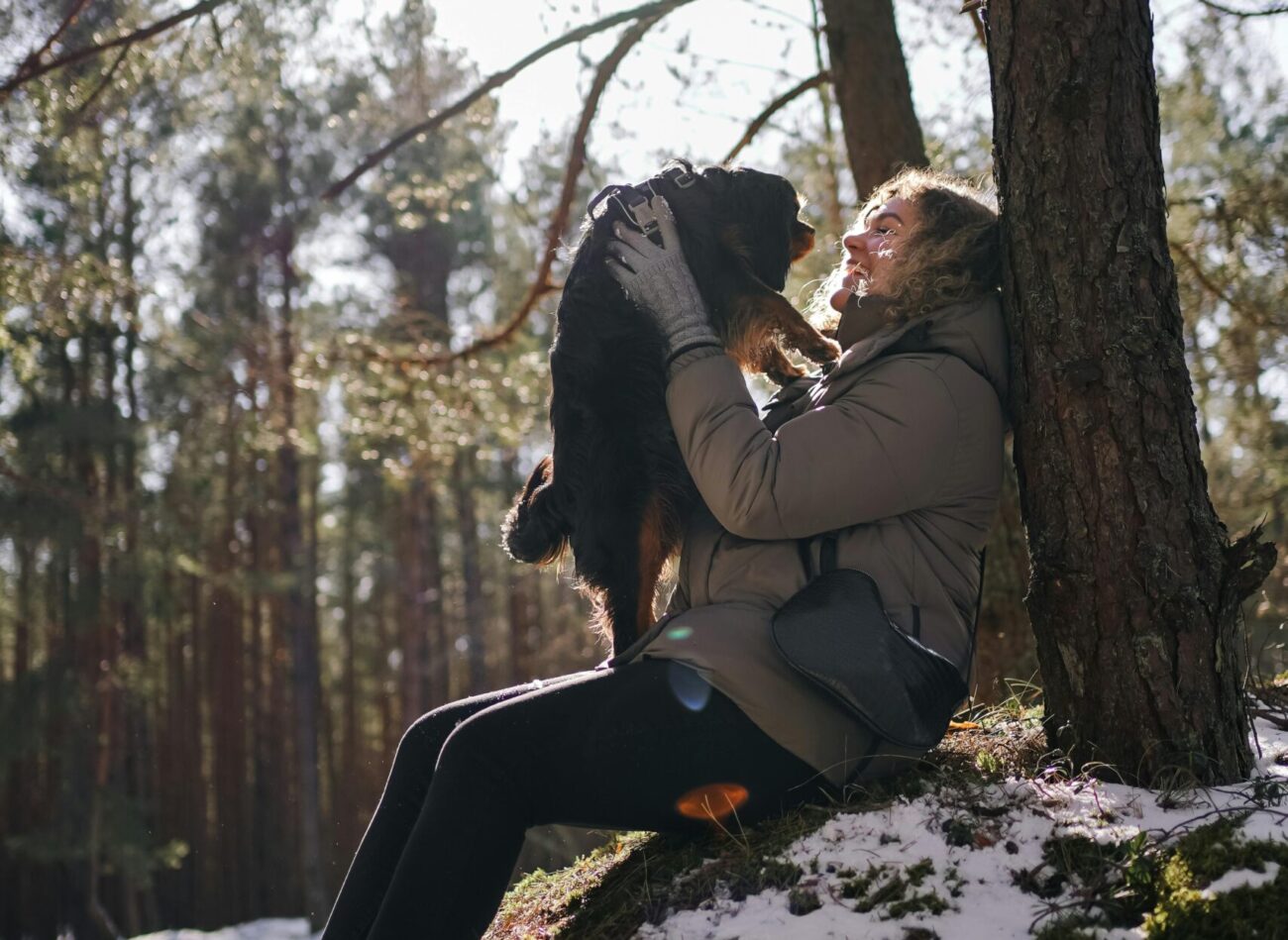 A joyful woman embraces her dog in the winter forest of Jūrmala, Latvia, basking in the warm sunlight.