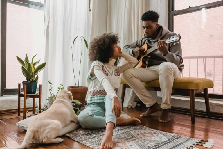Full body of black boyfriend playing acoustic guitar for African American girlfriend while spending time together in room with dog
