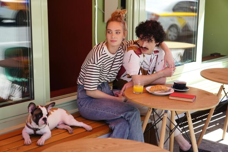 A couple sits outside a cafe with a dog, croissant, and coffee.