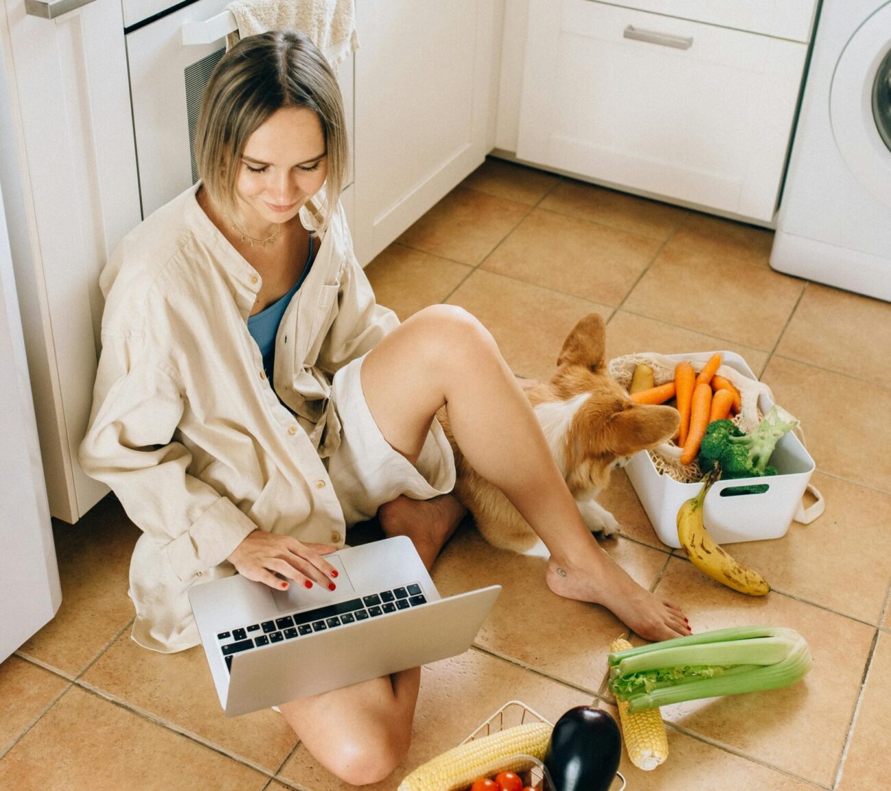 Woman sitting on kitchen floor with laptop, surrounded by fresh vegetables and groceries.
