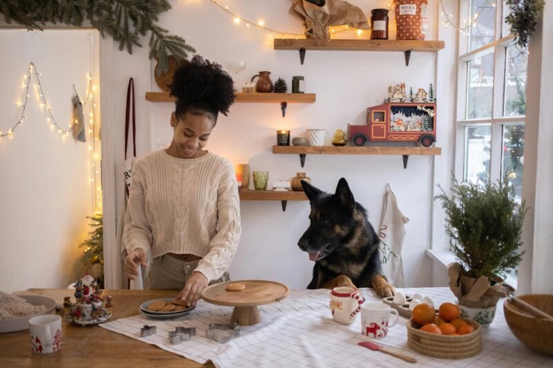 A joyful woman baking cookies with her German Shepherd in a festively decorated kitchen.